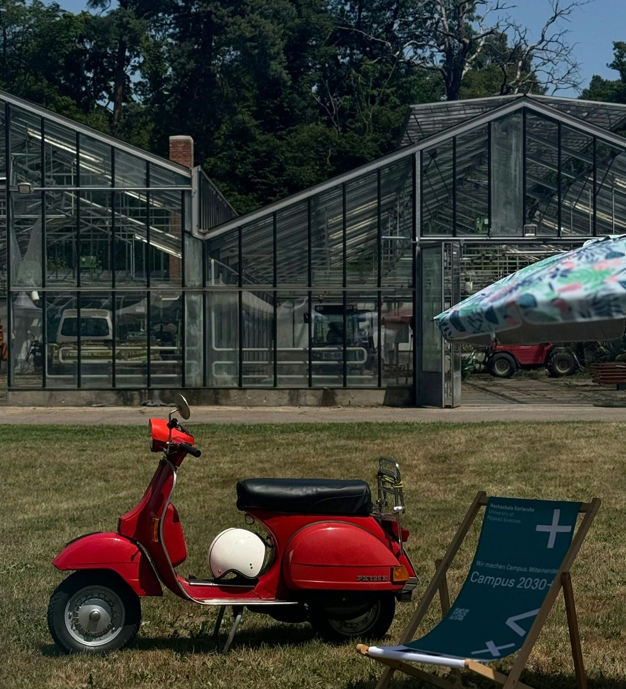 Rote Vespa und Liegestuhl vor Gewächshäusern in der Schlossgärtnerei Karlsruhe, umgeben von Grün und sommerlicher Stimmung 