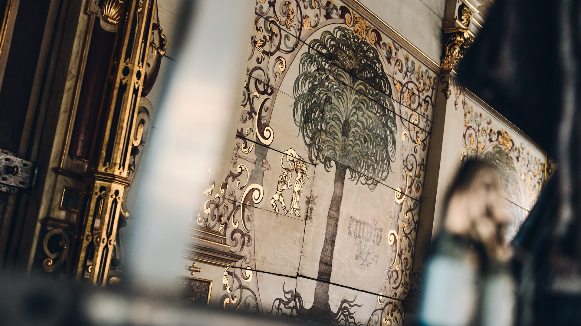 Im Vordergrund unscharfe Strukturen, im Hintergrund bemaltes Mauerwerk. Das Motiv: u.a. Ornamente und eine Palme.