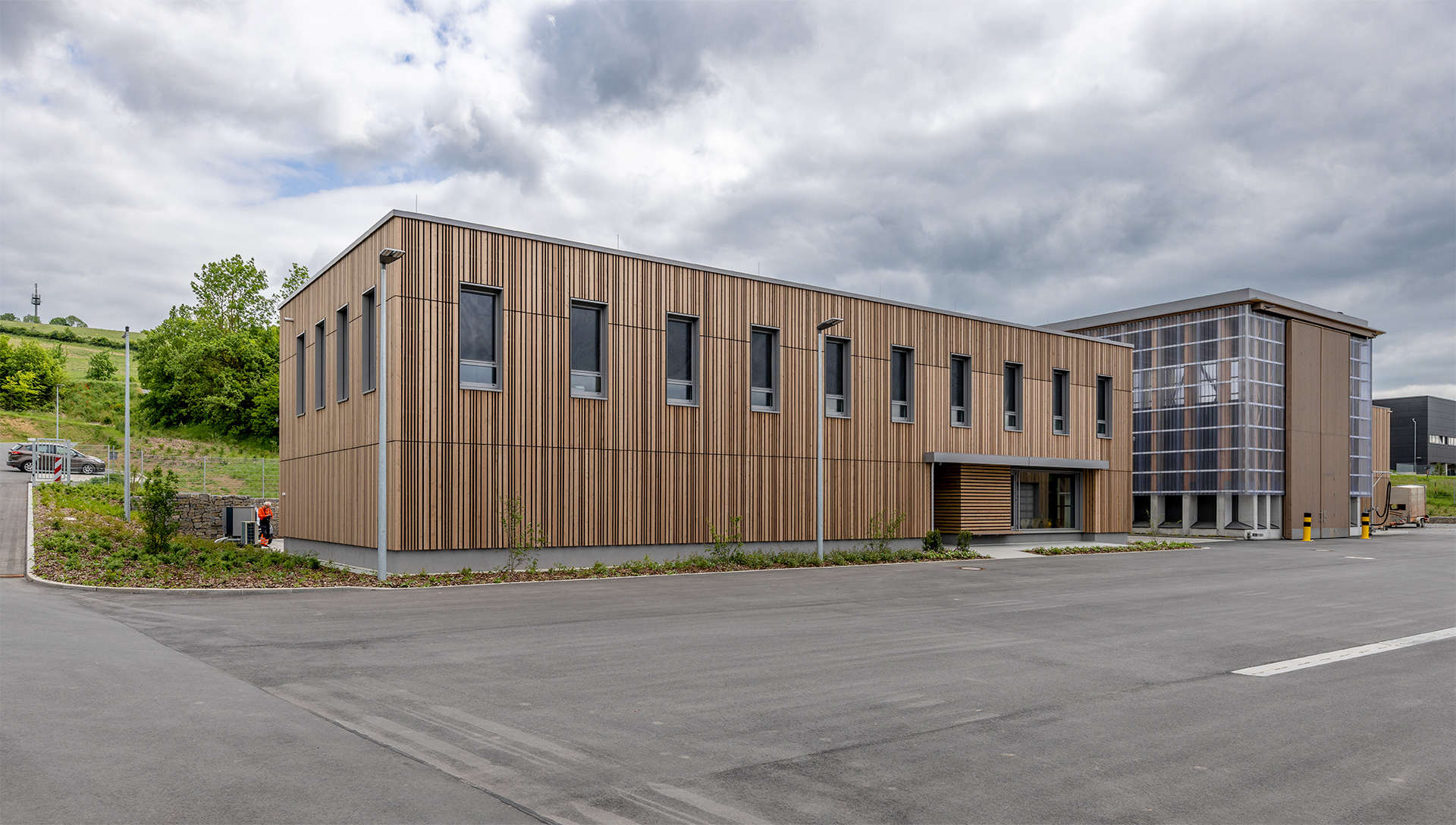 Zwei moderne Gebäude, die eine Fassade hauptsächlich aus Holz, die aus einem transparenten Material. Davor eine Asphalfläche.