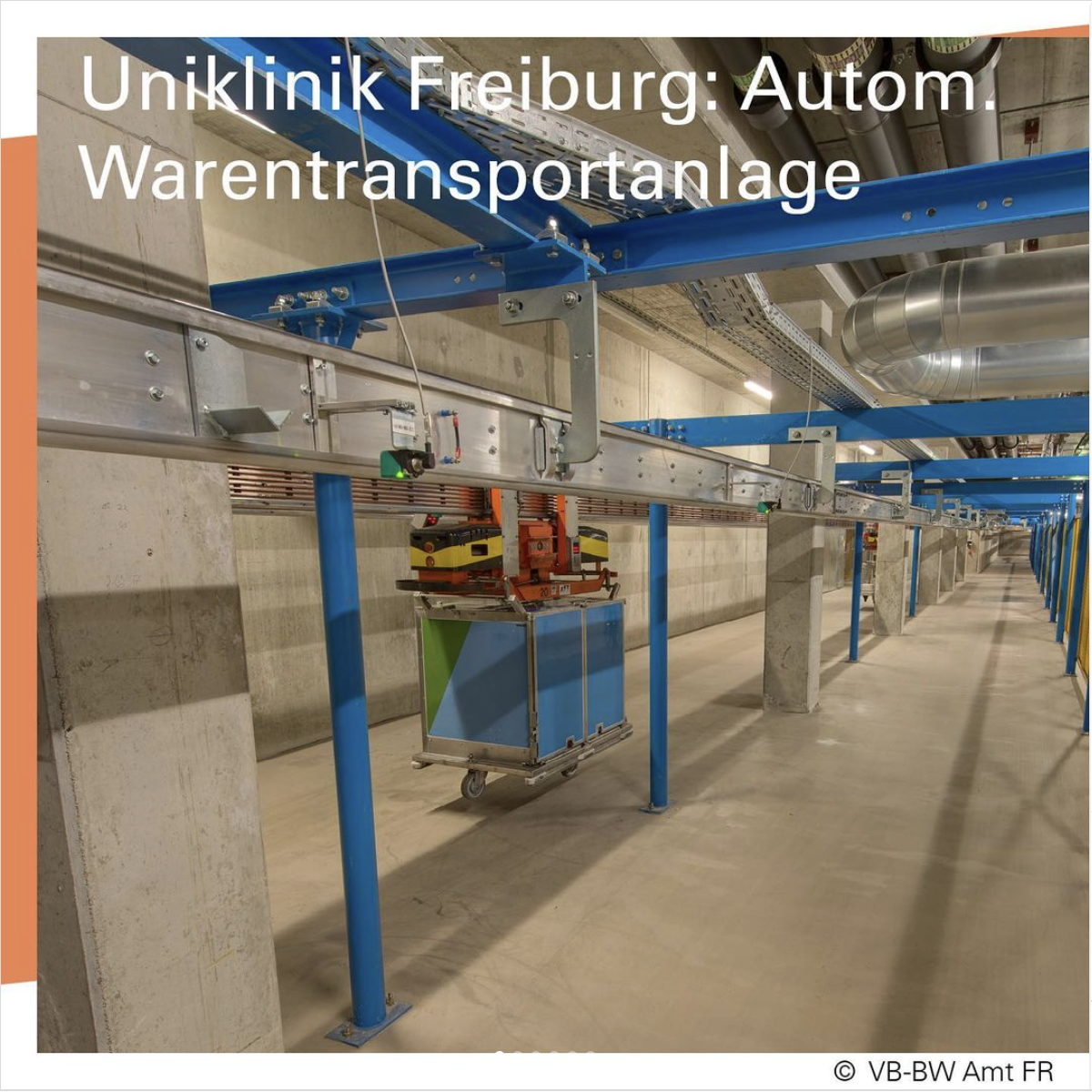 An einer Stahlträgerkonstruktion im inneren eines Gebäudes hängt eine blau-grüne Kiste auf Rollen.