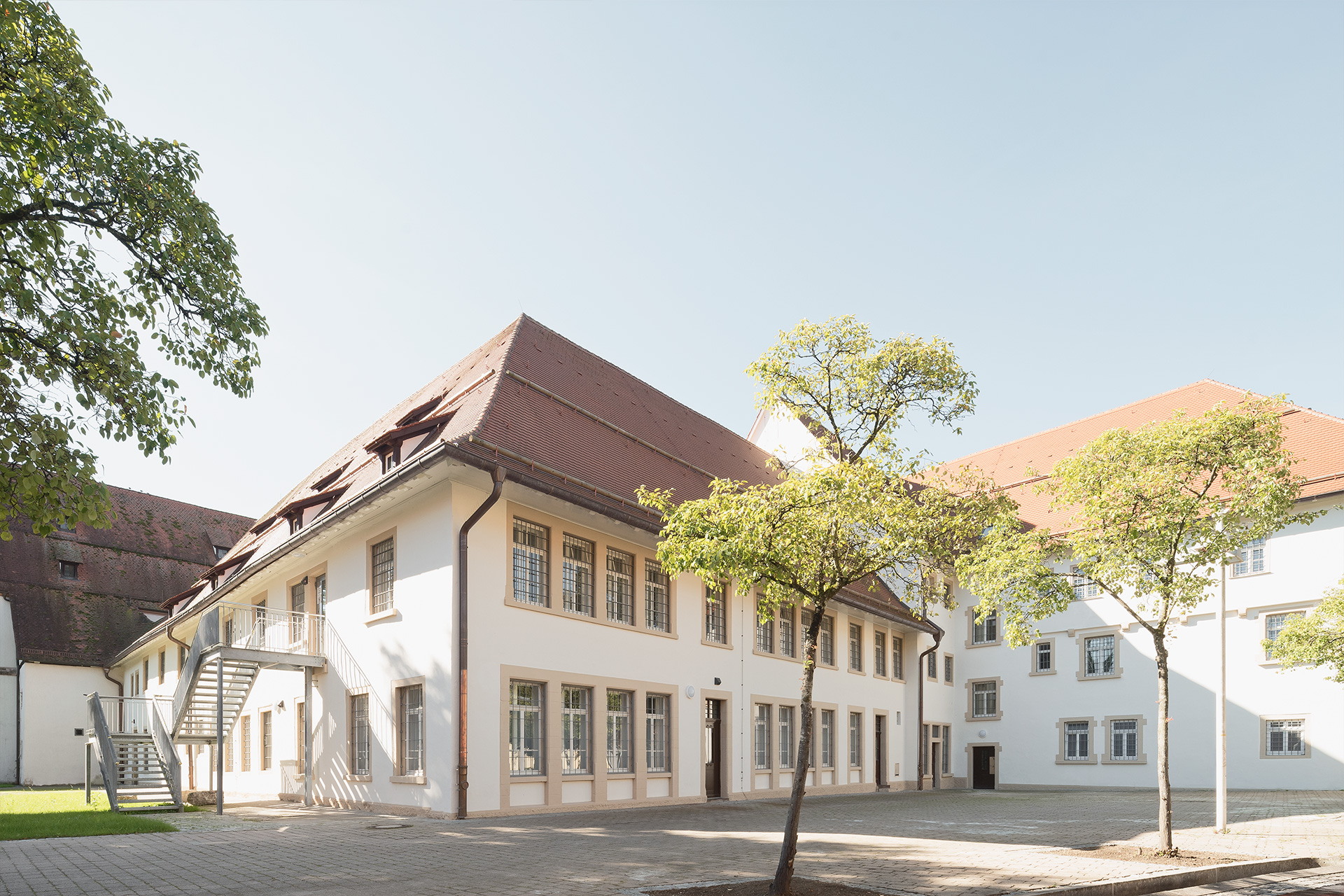 Ein Gebäude mit mehreren Trakten, vergitterten Fenstern und einer metallenen Außentreppe. Davor ein paar Bäume.