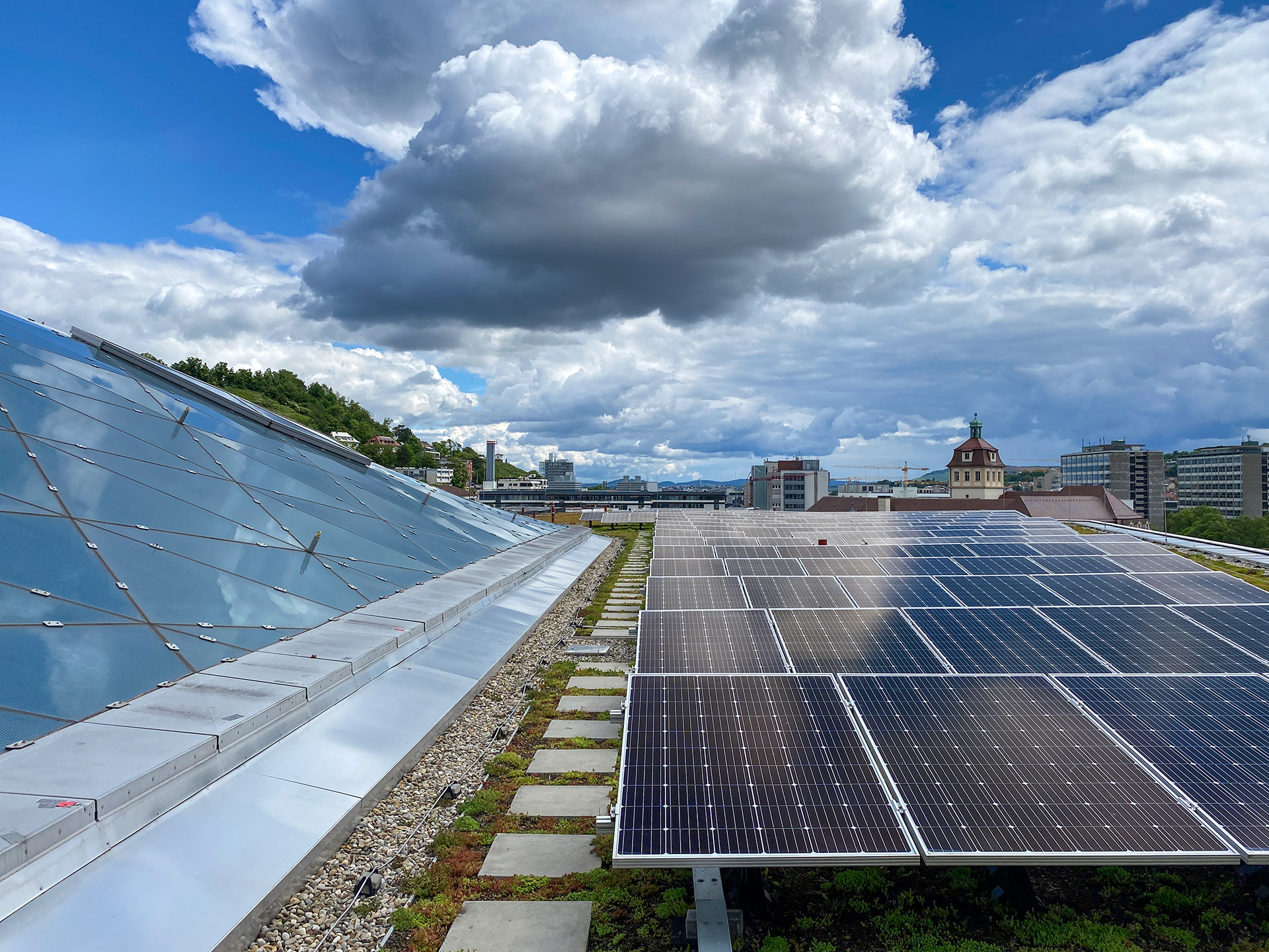 Links eine schräge, gläserne Dachfläche. Rechts eine flache, leicht begrünte Fläche, die mit PV-Modulen bestückt ist.