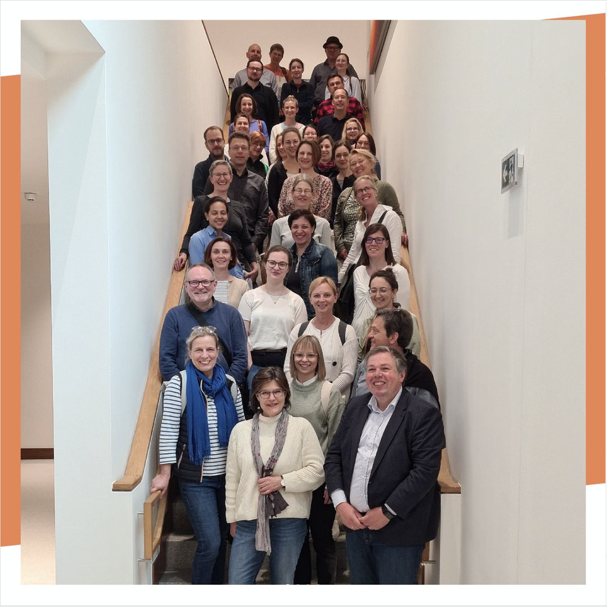 Gruppenbild: Auf einer relativ schmalen Treppe, eingefasst von weißen Wänden, stehen ca. 35 Personen.