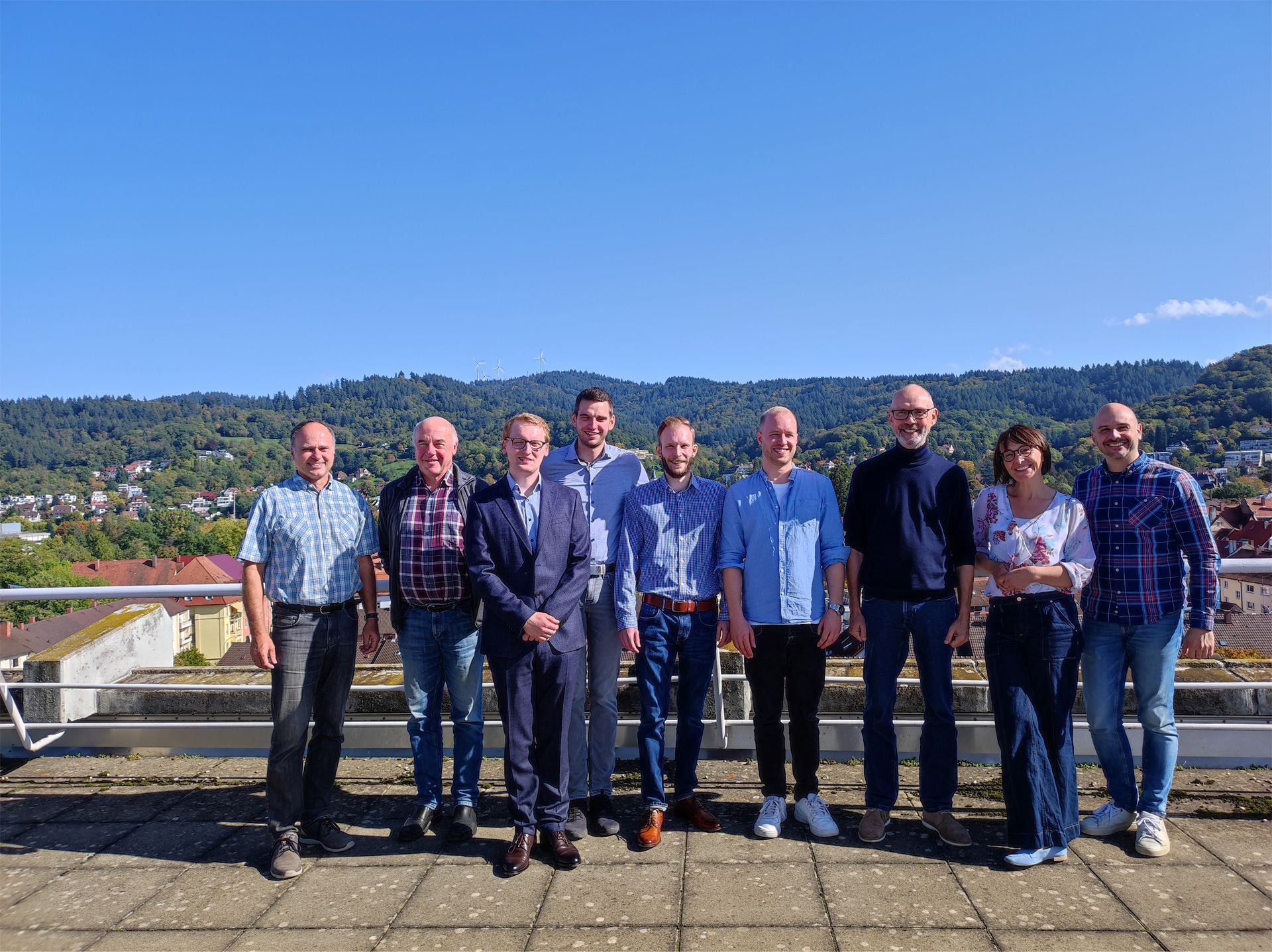 Gruppenbild: 9 Personen stehen auf einer Dachterrasse und blicken in die Kamera. Im Hintergrund eine bewaldete Stadt.