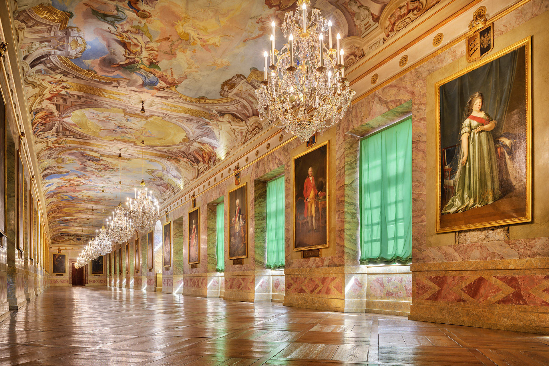 Ein Saal, üppiges Barock, mehrere große Kronleuchter und ein Gemälde an der Decke, Ganzkörper-Portraits an den Wänden.