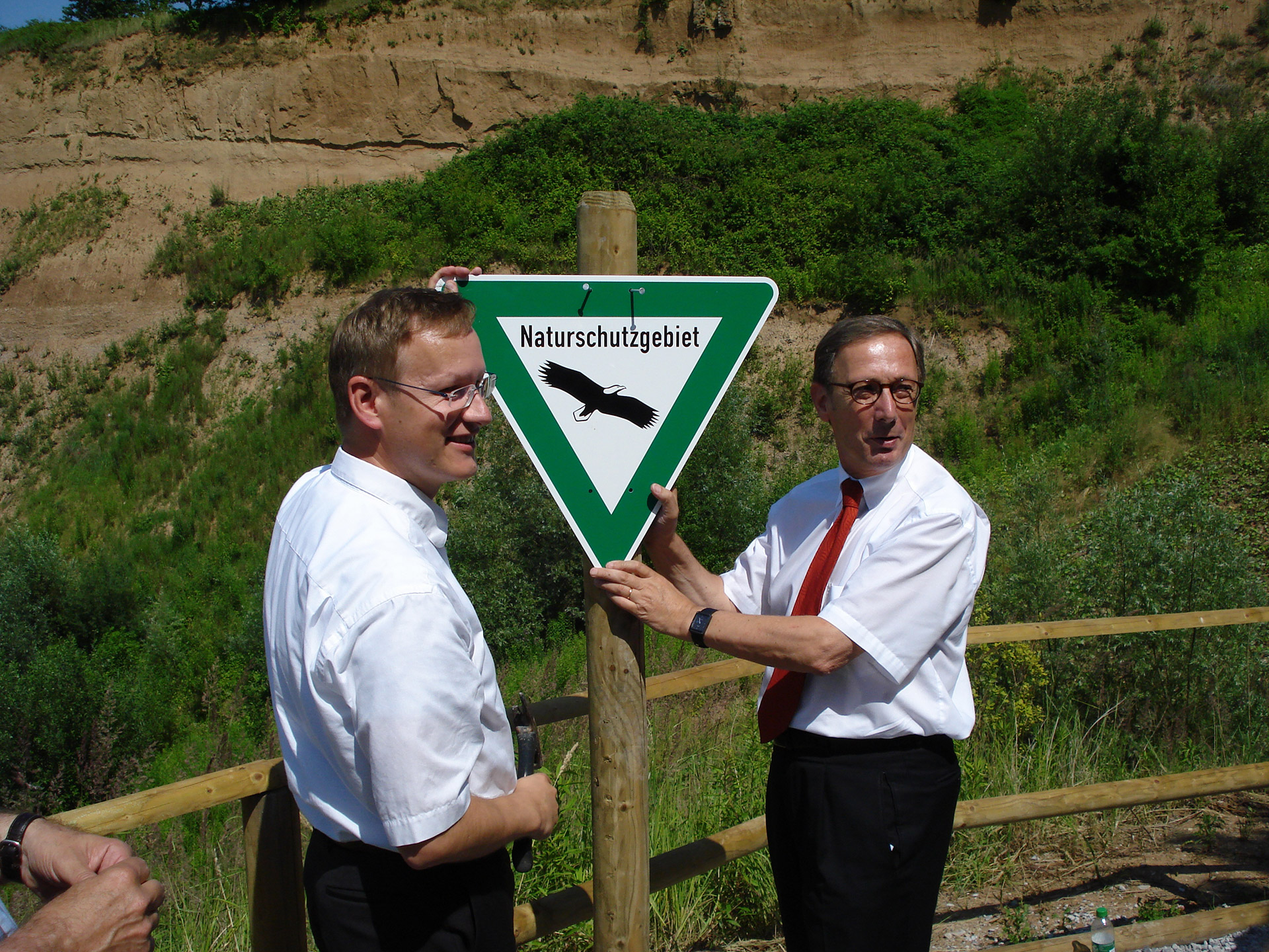Zwei Personen, in weißen Hemden und Krawatte, bringen an einem Holzpfahl ein Naturschutzgebiet-Schild an.