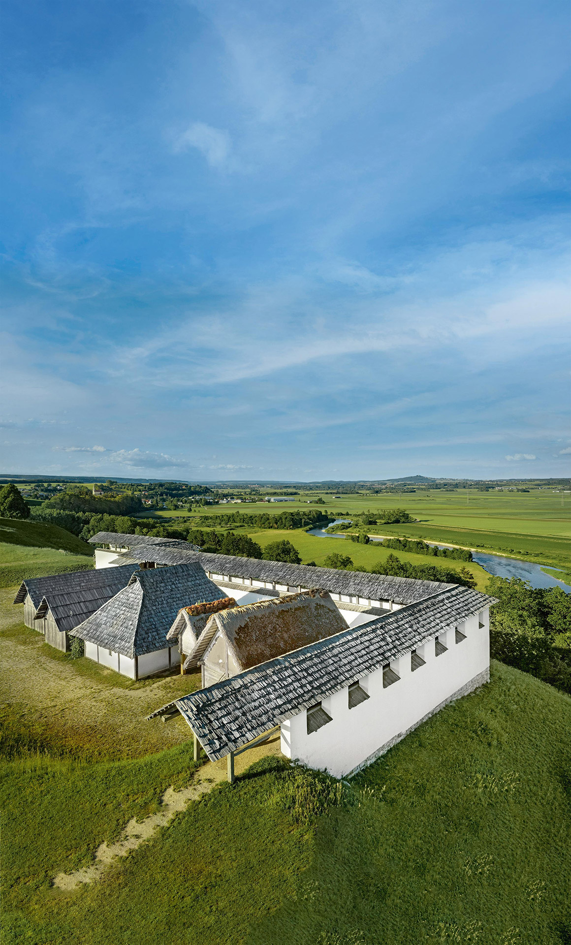 Blick in eine grüne Landschaft über historische Gebäude neben einer ums Eck gebauten Lehmziegel-Mauer hinweg.