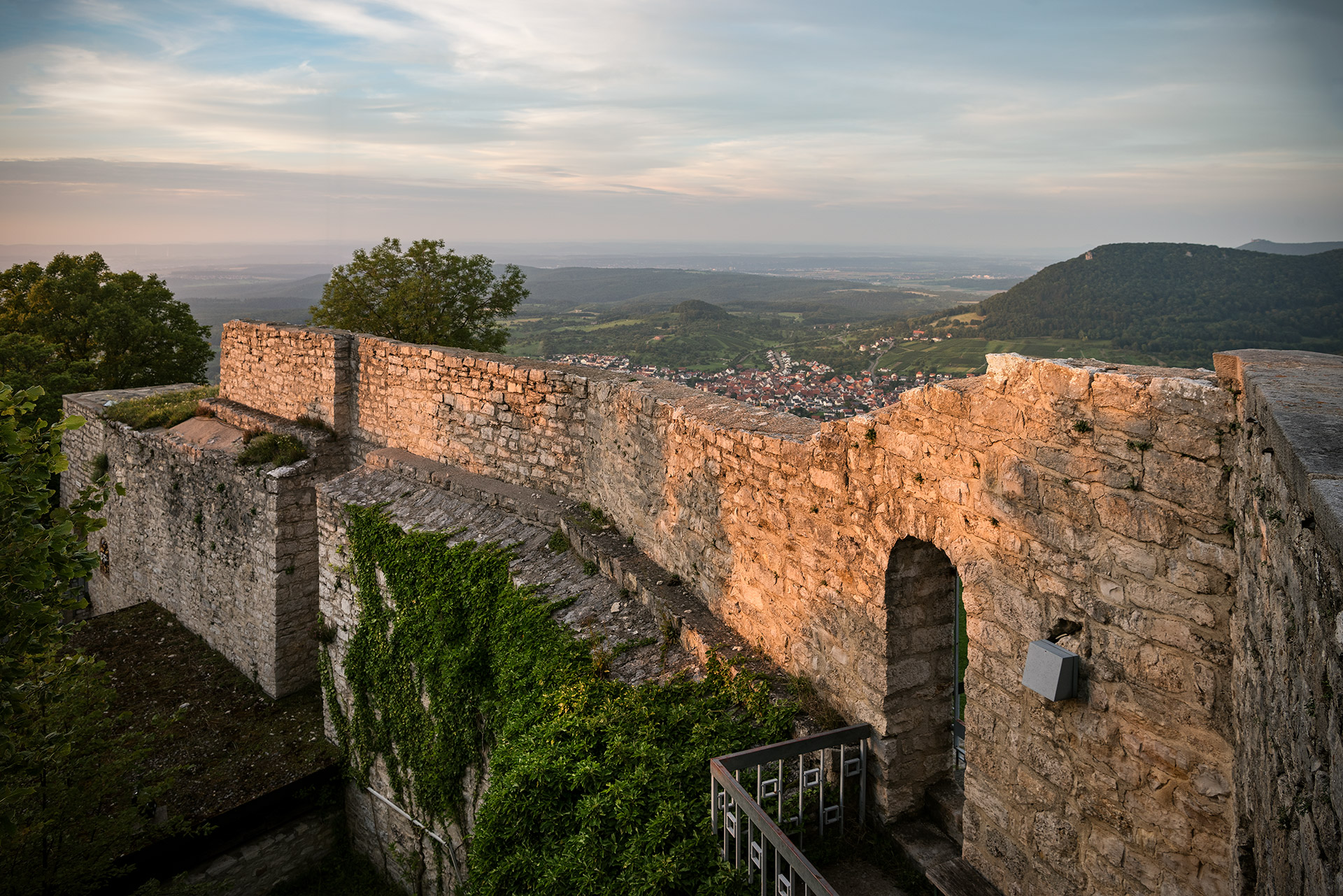Der Blick geht über eine von der tief stehenden Sonne angestrahlte, zum Teil grün bewachsene Ruinenmauer ins Weite