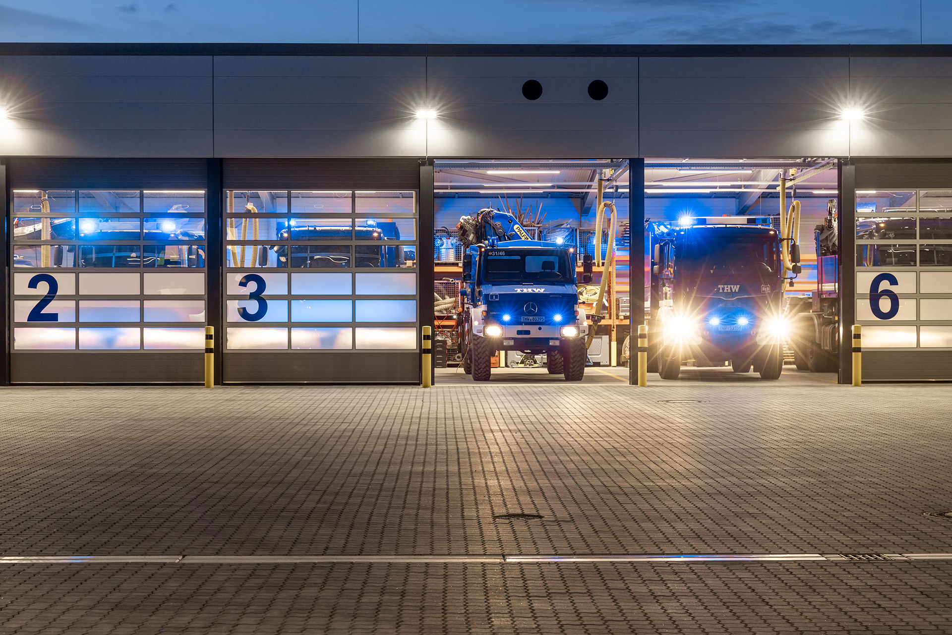 Eine beleuchtete Kfz-Halle des Technischen Hilfswerks in der Dämmerung, zwei Tore sind geöffnet und zeigen die LKWs darin.