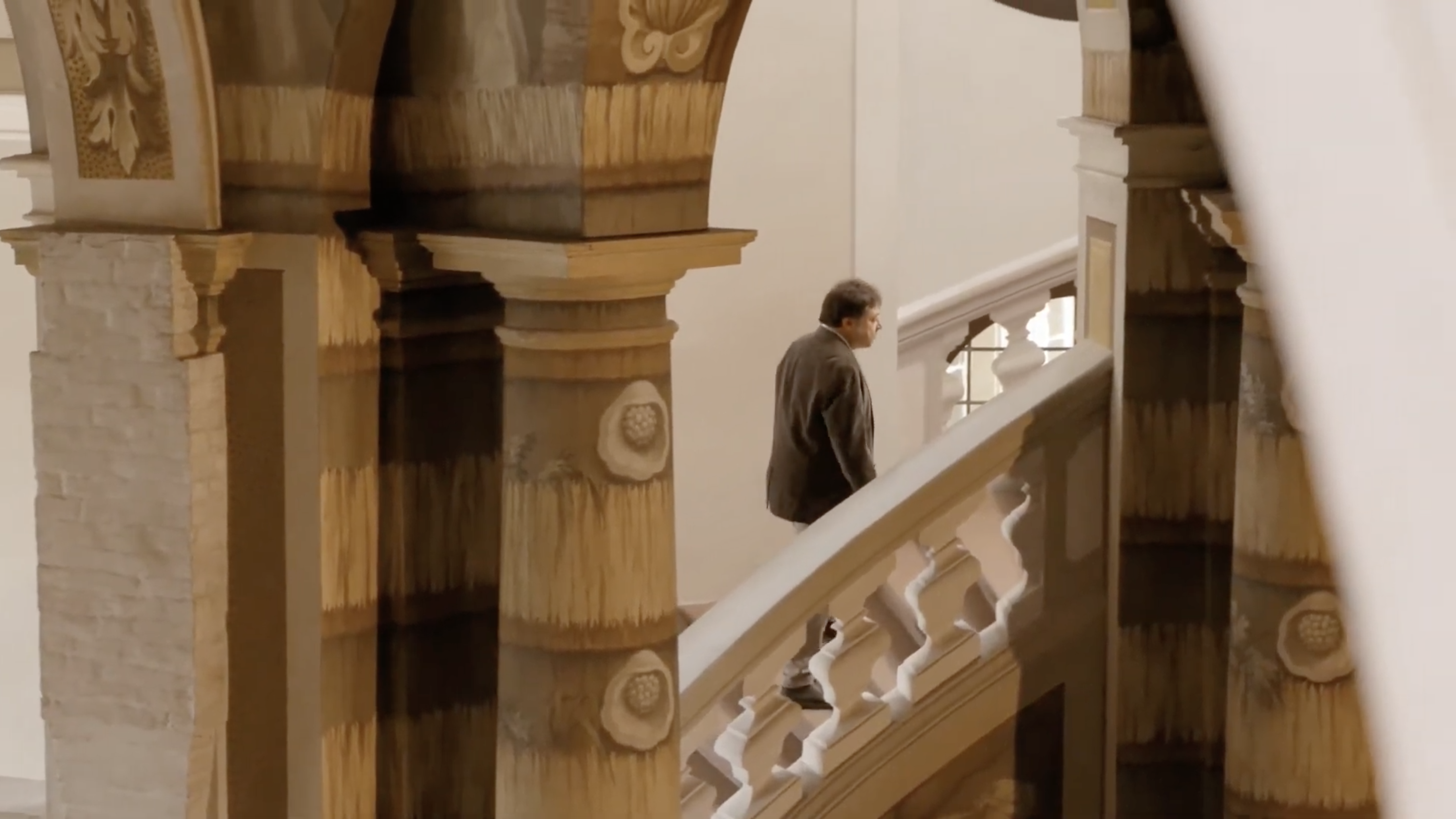 Im Vordergrund Säulen und im Hintergrund eine Treppe mit einem massiven Geländer aus Stein, die eine Person hochläuft.