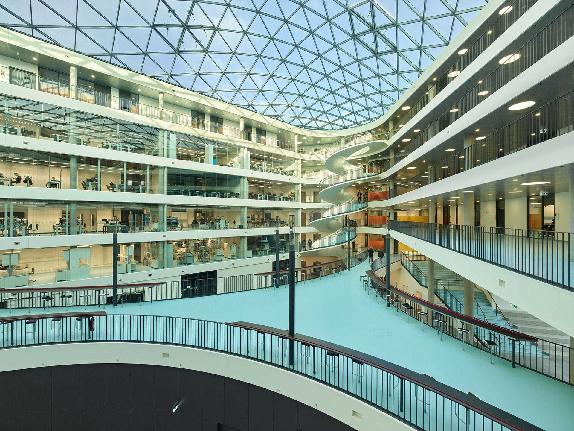 Ein großes Atrium mit gläserner Decke und spiralförmiger Wendeltreppe. Verglaste Wände zeigen die Räumlichkeiten. 