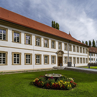 Ein zweigeschossiges historisches Gebäude mit umrahmten Fenstern. Davor eine Rasenfläche und ein blumenumrahmter Brunnen.