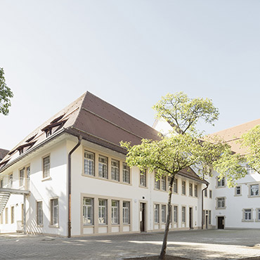 Ein großes, mehrgliedriges Gebäude mit vergitterten Fenstern und metallener Außentreppe. Davor ein Baum.