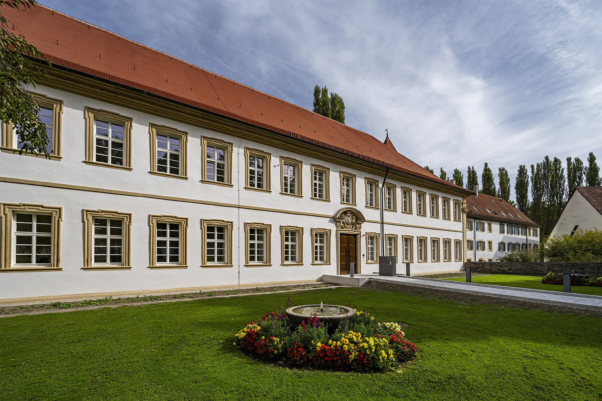 Ein zweigeschossiges historisches Gebäude mit umrahmten Fenstern. Davor eine Rasenfläche und ein blumenumrahmter Brunnen.