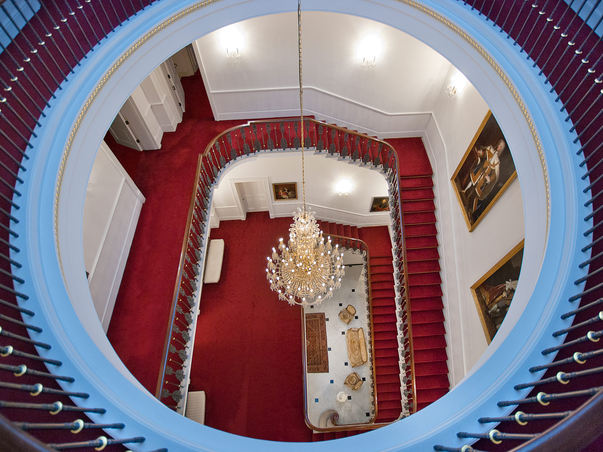 Das Treppenhaus von oben nach unten fotografiert: weiße Wände, roter Teppich, auffällig gestaltete Geländer. 