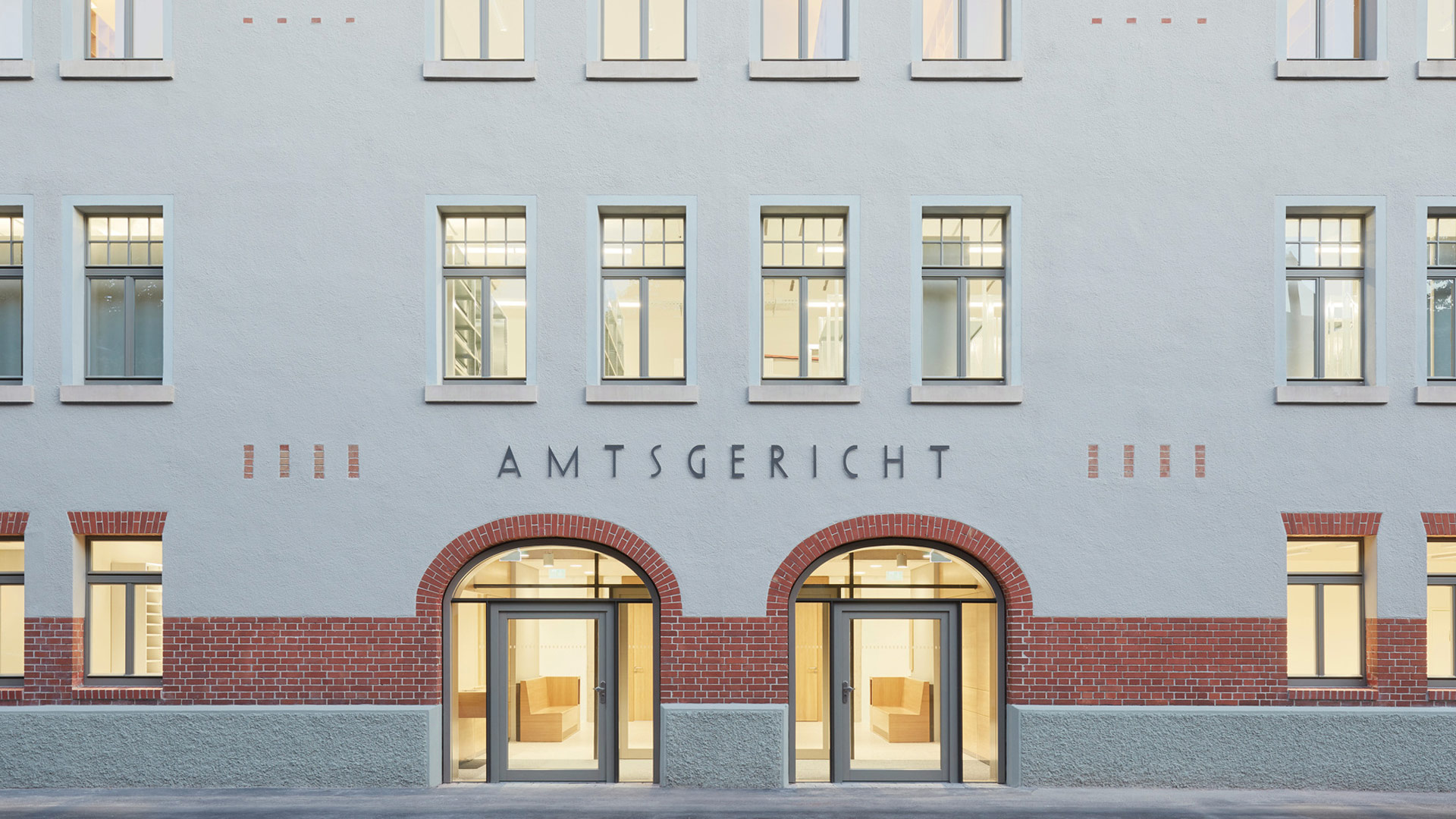 Ausschnitt einer grauen Fassade mit zum Teil rot abgesetzten Fenstern. Über den beiden Türen steht: „Amtsgericht“.