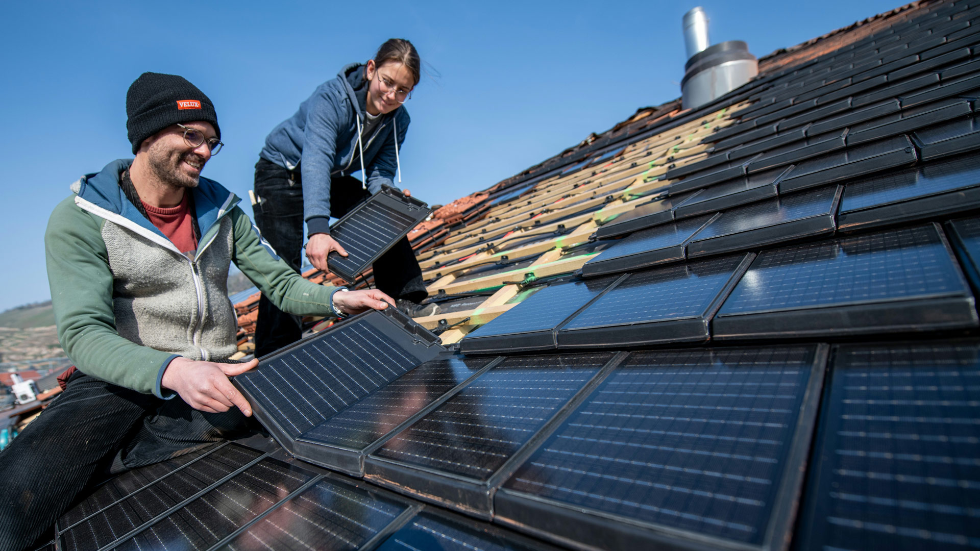 Zwei Personen befinden sich auf einem halb gedeckten Dach. Eine bringt Solardachziegel an, die andere reicht ihr die Ziegel an.