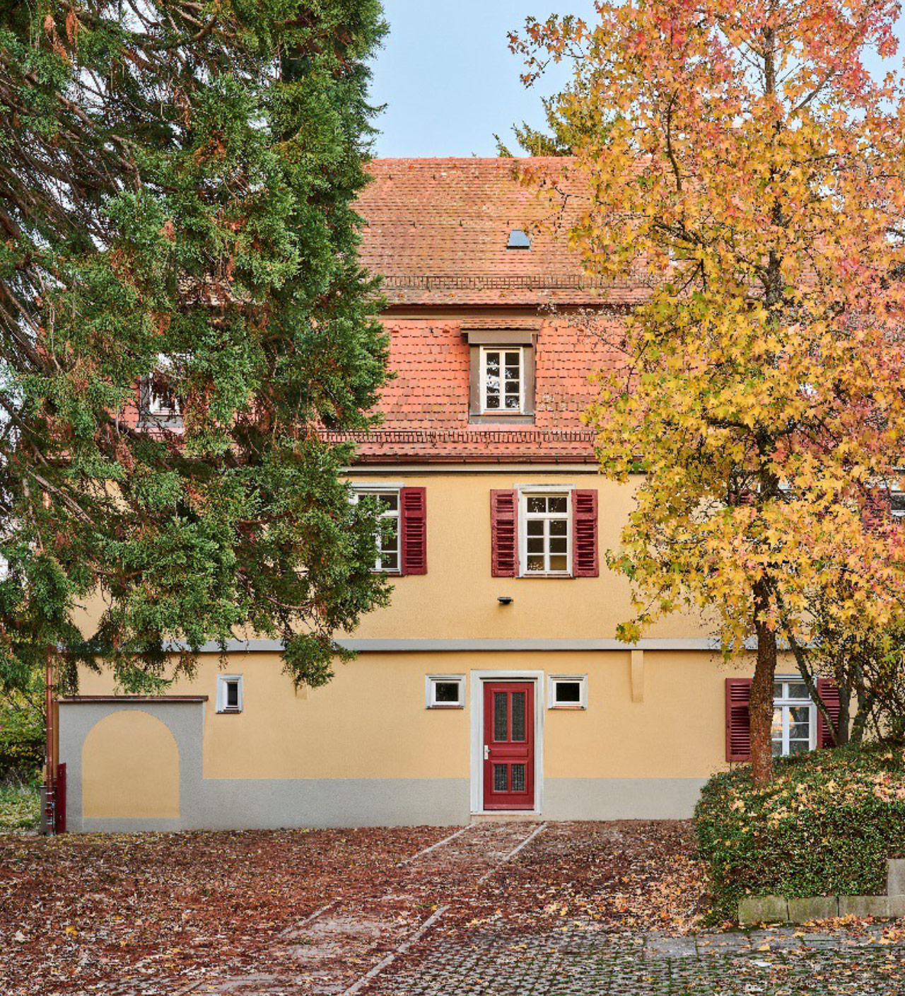 Straßenansicht des sanierten evangelischen Pfarrhauses in Stuttgart-Plieningen: zweigeschossiger Putzbau aus dem 18. Jahrhundert mit rotem Ziegeldach, dunkelroten Fensterläden und Eingangstür, flankiert von Laubbäumen.