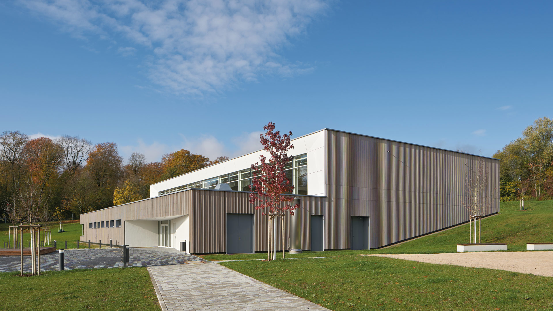 Ein Gebäude mit zweiteiligem, terrassiertem Baukörper, umgeben von einer Grünanlage mit Jungbäumen, Rasen und Kiesflächen.