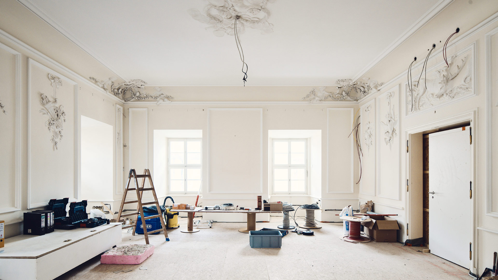 Baustellen-Atmosphäre in einem historischen Raum mit Stuck an Wänden und Decke. Elektrokabel ragen aus Decke und Wänden, diverse Handwerkerutensilien sind verteilt.